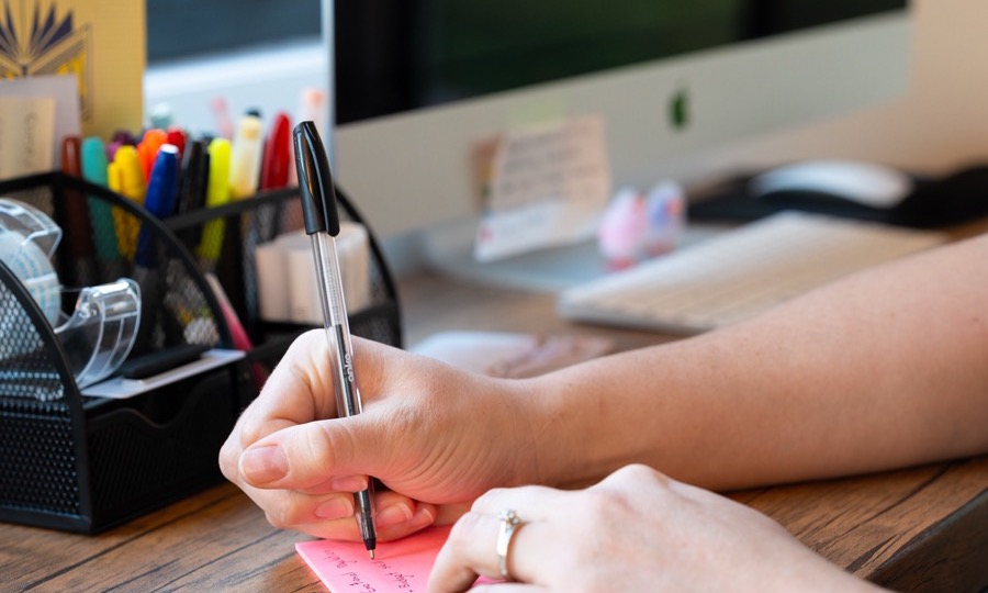 Hands writing a note at a desk beside a jar of pens