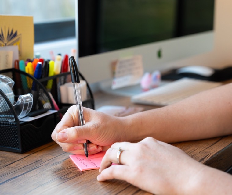 Hands writing a note at a desk beside a jar of pens